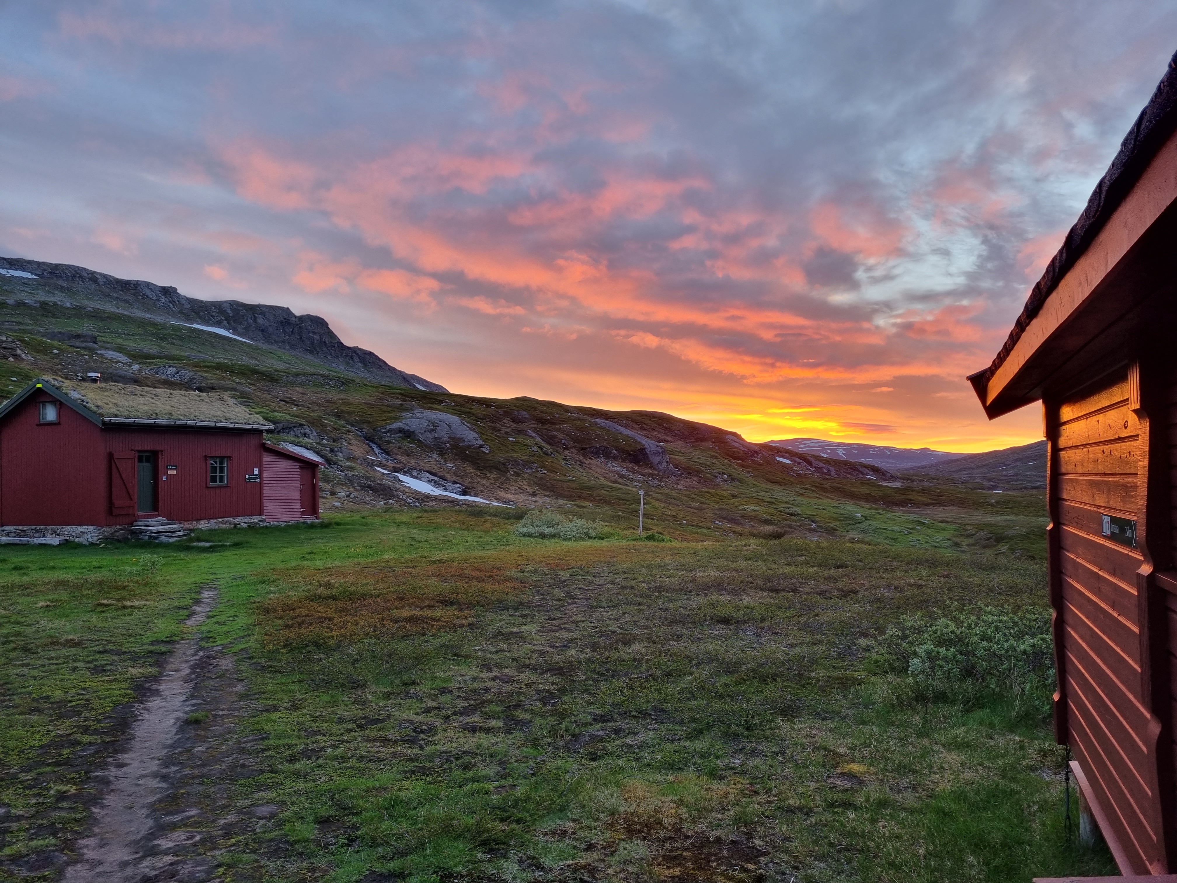 En rødbrun hyttevegg og ei rødbrun hytte i bakgrunnen. Det er lyng og fjell. En lys sommernatt med gul og rød himmel.